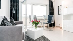 A modern living room with a grey sofa, a white coffee table with a vase of flowers, a desk with a chair, and a view of the city through large windows. - Modern vacation apartment with sauna, solarium and heated indoor pool in Westerland (Westerland)