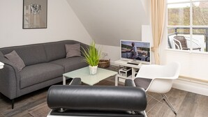 Un saló modern amb un sofà gris, una cadira de cuir negre, una taula petita amb una planta i una televisió que mostra un home treballant.