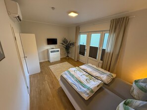 Un dormitorio con cama, televisor, escritorio, planta en maceta y ventana con cortinas.
