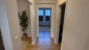 Un couloir doté d’un plancher en bois, de murs blancs, d’une plante en pot et d’une porte donnant sur une pièce avec une fenêtre.