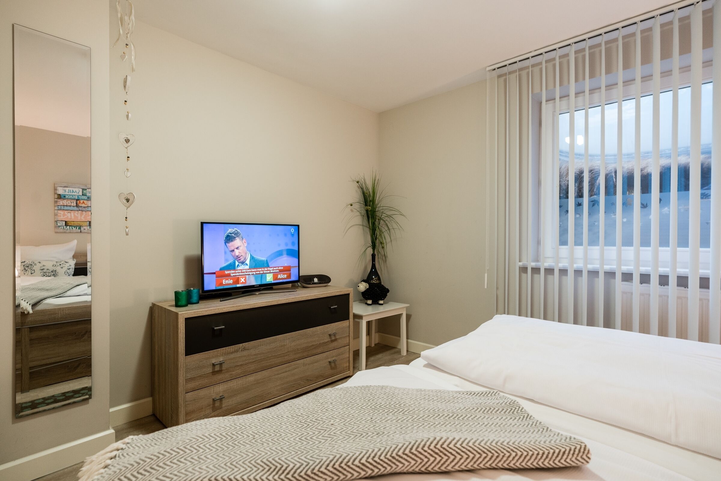 Hotelkamer met een bed, een televisie op een houten dressoir, een tafeltje met een plant en een raam met zonwering.