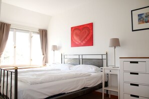 A bedroom with a bed, bedside tables, a nightstand, a dresser, a window with curtains, and a red heart artwork on the wall.