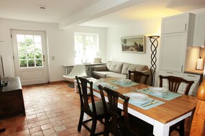 A dining area with a wooden table set for four, surrounded by chairs. There is a window with a view of greenery outside, a sofa in the background, and a cabinet to the right.