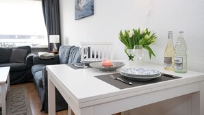 Ein modernes, helles Zimmer mit einem Essbereich, der einen weißen Tisch, Teller und eine Vase mit Blumen zeigt. Im Hintergrund sind eine Couch und eine Lampe zu sehen.