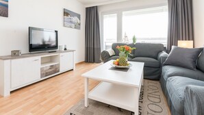Ein modernes Wohnzimmer mit Fernseher, einem weißen Couchtisch mit Obst, einem grauen Sofa und Meerblick durch große Fenster.