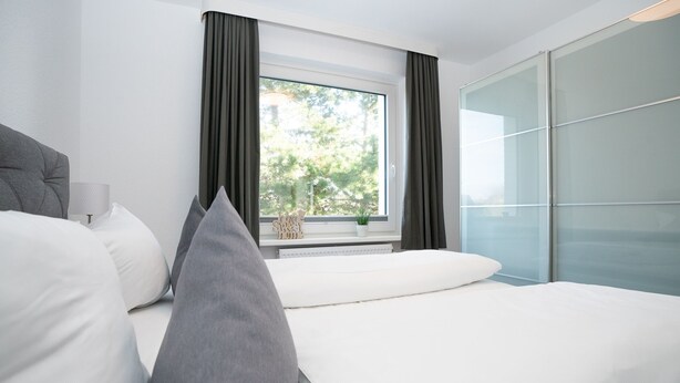 Ein modernes Hotelzimmer mit einem großen Bett, einem Fenster mit Vorhängen, Blick auf Bäume und einer Trennwand aus Glas.