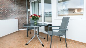 A balcony with a round table and a chair, a vase of red flowers, and a view of the city through a window. - Comfortable 2-room vacation apartment in the Nordmarkhof with balcony in Westerland (Westerland)