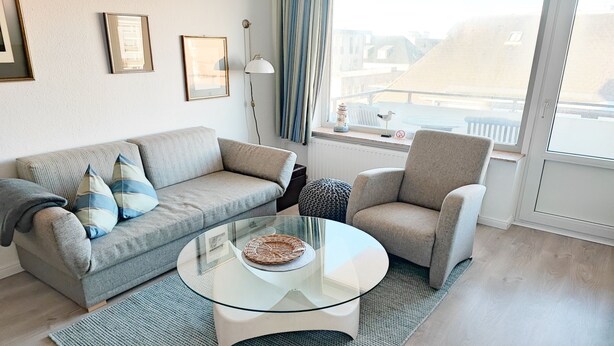 A modern living room with a glass coffee table, a sofa with striped cushions, a chair, and a view of the city through large windows. - Comfortable vacation apartment with balcony in Westerland (Westerland)