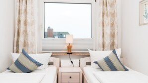 A hotel room with two beds, a nightstand, a lamp, and a window with a view of buildings. - Comfortable vacation apartment with balcony in Westerland (Westerland)