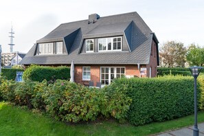 Une maison à deux étages, avec un toit en pente, une façade en briques et un jardin bien entretenu agrémenté de haies et d’une pelouse.