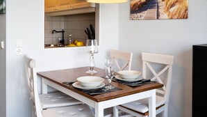 Un comedor con una mesa de madera dispuesta para dos personas, sillas blancas y vista a la cocina al fondo.