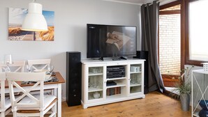 Una sala de estar moderna con un televisor de pantalla plana sobre un mueble blanco, una mesa de comedor con sillas, y un ventanal con cortinas.
