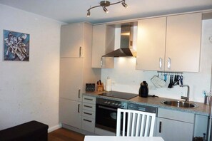 Eine kleine Küche mit Herd, Backofen und Spüle. Ein weißer Stuhl und ein schwarzer Tisch sind vorhanden.