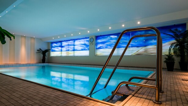 An indoor swimming pool with clear blue water, a metal ladder, tiled floor, and a large window with a sky view. - Modern vacation apartment with sauna, solarium and heated indoor pool in Westerland (Westerland)