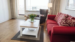Uma sala de estar moderna com um sofá vermelho, uma poltrona cinza e uma mesa de centro branca. Há uma janela com cortinas, um vaso com flores e uma luminária em uma mesinha lateral.