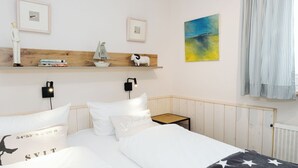 Quarto de hotel com cabeceira de madeira, cama com roupa de cama branca, mesa de cabeceira pequena e prateleira com objetos decorativos.