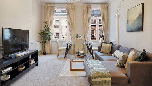 A modern living room with a flat-screen TV, a sofa, a coffee table, and a dining area with a glass table and chairs. - Hertford Flat 12 (London)