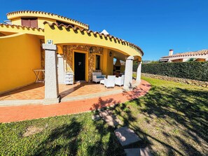 Eine gelbe Villa mit Steinaußenwand, einer Terrasse mit Gartenmöbeln und einer Mauer, die das Grundstück umgibt.