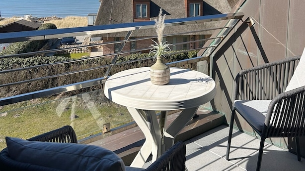 A balcony with a table and chairs, overlooking a coastal area with a thatched-roof building and a view of the sea. - Exclusive vacation apartment with a view of the sea (Wenningstedt)