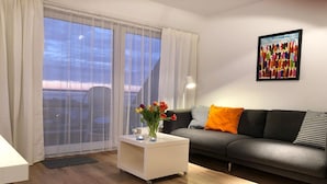 A modern living room with a grey sofa, a white side table with a vase of flowers, and a large window with sheer curtains. - Exclusive vacation apartment with a view of the sea (Wenningstedt)