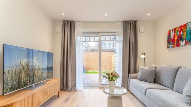 A modern living room with a flat-screen TV, grey sofa, and a small round table with a vase of flowers. - This beautiful and new terraced house is centrally located in Westerland (Westerland)