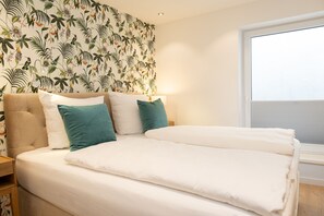 A bedroom with a large bed, a floral wallpaper, and a window with blinds. - Exclusive thatched house in Wenningstedt with sauna, garden and modern furnishings (Wenningstedt)