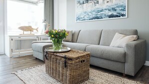 Ein Wohnzimmer mit grauem Sofa, einem Korbtisch mit Blumenvase, einem Gemälde an der Wand und einem Fenster mit Blick auf ein Gebäude.
