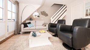 Una sala moderna con un sofá, un sillón de cuero negro, una mesa de centro con frutas, una lámpara de pie y una escalera.