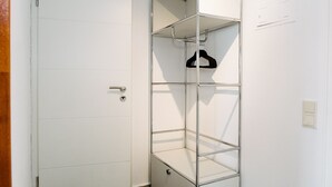 Un armario blanco con repisas metálicas y una barra para colgar, una puerta con manija y un interruptor de luz en la pared.