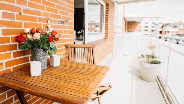 Una mesa de madera con sillas y un jarrón de flores en un balcón con un muro de ladrillo y vistas a otros edificios.