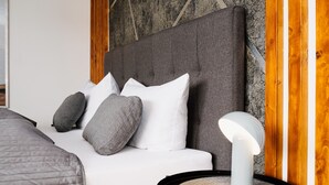 Un dormitorio moderno con un cabecero tapizado gris, almohadas blancas y grises, una mesita de noche redonda con una lámpara blanca y un posavasos tejido.