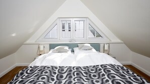 Een slaapkamer met een schuine kap, een bed met witte beddengoed, een patroon deken en een raam met witte luiken.