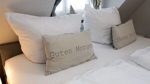 Una cama bien hecha con dos almohadas que dicen "Guten Morgen" y una ventana con cortinas.
