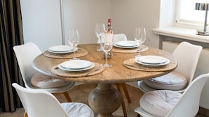 Una mesa de comedor redonda de madera con sillas blancas acolchadas, servida con platos, vasos y una botella de vino.