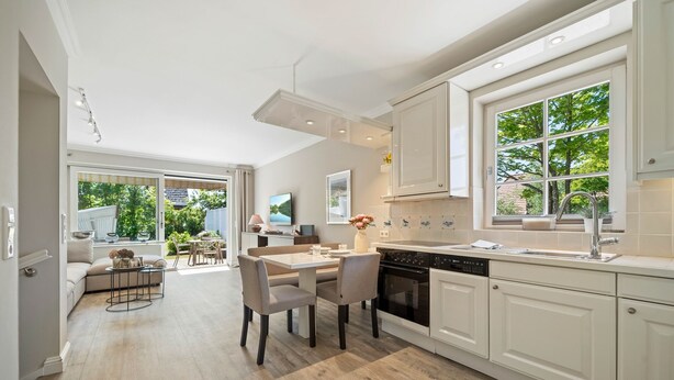 Een moderne keuken met witte kasten, een eethoek met een tafel en stoelen, en uitzicht op het groen door grote ramen.