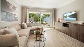 Een moderne woonkamer met een beige bank, een bijzettafeltje met decoratieve items, een flatscreen televisie op een houten kast en een groot raam met uitzicht op groen en een gebouw.