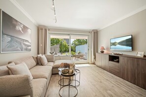 Ein modernes Wohnzimmer mit einem beigen Sofa, einem Couchtisch mit Dekoration, einem Flachbildfernseher auf einer Holzkommode und einem großen Fenster mit Blick auf Grünflächen und ein Gebäude.