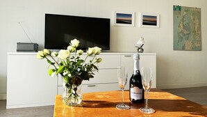 Et værelse med et træbord, en vase med hvide blomster, en champagneflaske og to glas. Et fladskærms-tv er monteret på væggen over et hvidt skab.