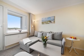 Ein modernes Wohnzimmer mit grauem Sofa, einem Couchtisch mit Blumenvase, einem kleinen runden Tisch mit Obst und einem Strandbild an der Wand.