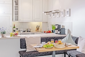A modern kitchen with white cabinets, a dining area with a bench, and a breakfast setup including fruits, bread, and a coffee maker. - Modern vacation apartment with sauna and heated indoor pool in Westerland (Westerland)