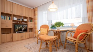A modern living room with wooden cabinets, a television, wicker chairs, a table, and a large window with curtains. - Comfortable apartment with balcony, elevator and parking space in Westerland (Westerland)