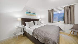 Une chambre à coucher avec un lit, des tables de chevet, une fenêtre avec des rideaux, une chaise et un tableau accroché au mur.