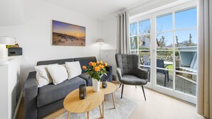 Un salon moderne avec un canapé, une table basse, un fauteuil, un tableau et une vue dégagée sur l’extérieur grâce à de grandes fenêtres.