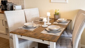 Mesa de jantar posta com pratos, tigelas e velas brancas, rodeada por cadeiras de vime com almofadas cinzentas.