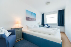 A hotel room with a bed, a chair, a nightstand, a lamp, a window with curtains, and a painting on the wall. - Comfortable vacation apartment with balcony in Haus Ankerlicht (Westerland)