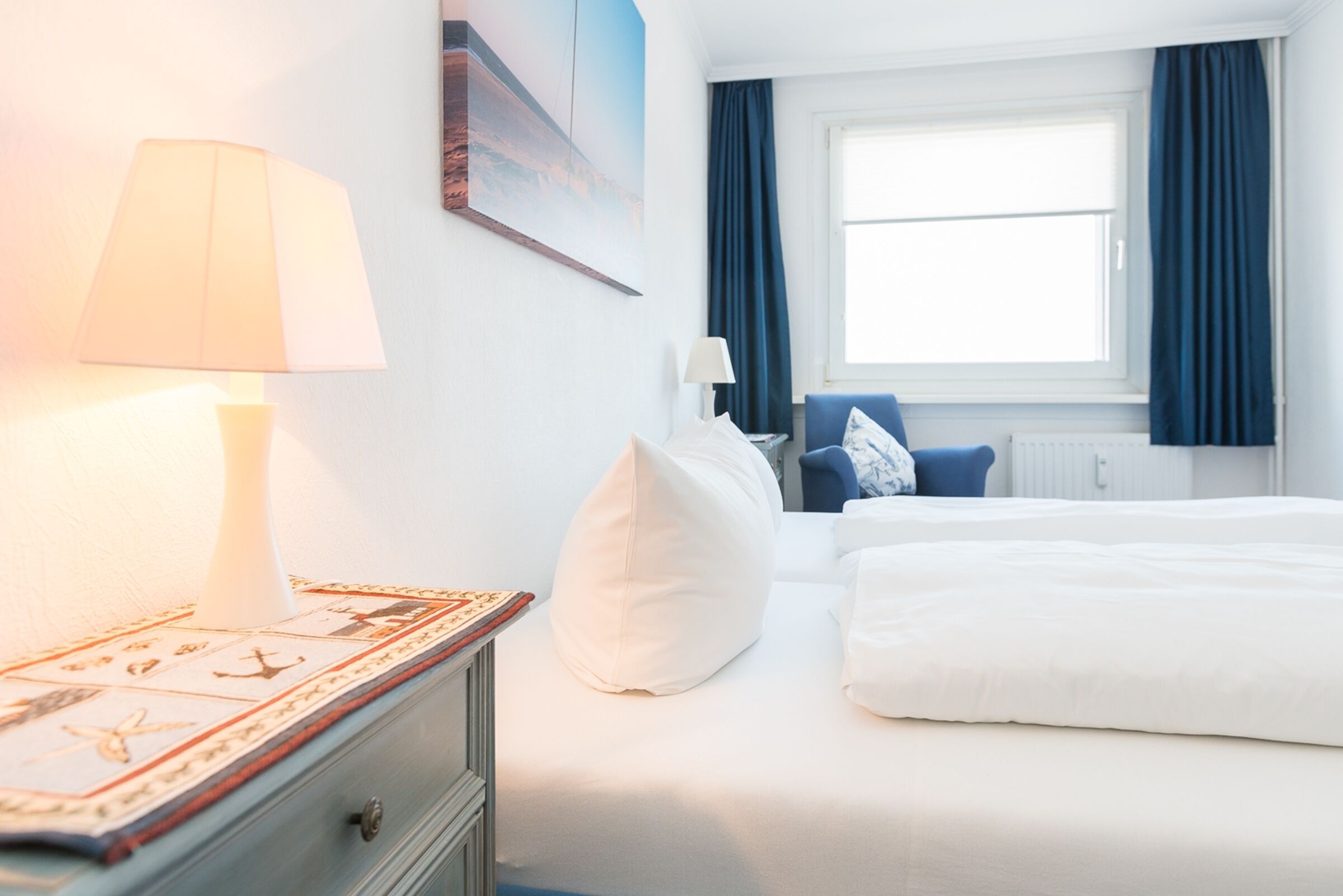 A hotel room with a bed, a nightstand with a lamp, a window with curtains, and a painting on the wall.