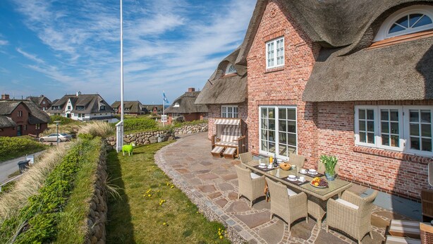Ein Haus mit Strohdach, Backsteinwänden und einem Kiesweg, der zu einer Terrasse mit Gartenmöbeln führt.