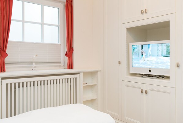 A bedroom with a bed, a window with curtains, a TV on a stand, and a white wardrobe.