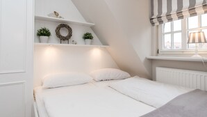 Et soveværelse med skråvæg, hvide vægge, en seng med hvide sengetøj, et vindue med stribet gardin og reoler med plantepotteplanter.