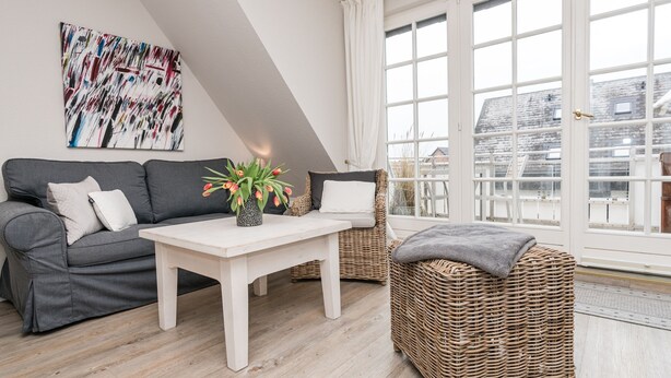 En moderne stue med en grå sofa, et hvidt sofabord og fletningsmøbler. Et stort vindue giver udsigt til bygninger udefra.